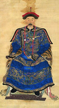 ภาพถ่ายขาวดำเต็มหน้าของชายคนหนึ่งที่กำลังนั่ง ใบหน้าซูบผอม สวมเสื้อคลุมที่ประดับด้วยลวดลายเมฆและมังกรอันซับซ้อน