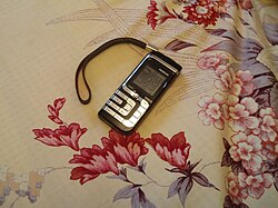 โทรศัพท์มือถือ Nokia 7260 สีดำ ปุ่มกดสีเงิน และสายคล้องข้อมือสีดำ วางอยู่บนผ้าปูโต๊ะลายดอกไม้