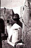 Black-and-white photograph showing an open-air scene of ruins with a white, peaked centotaph in between two short white columns
