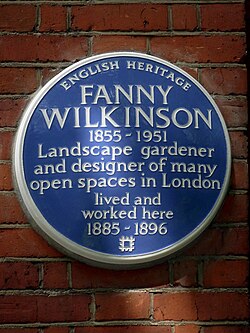 ป้ายกลมสีน้ำเงิน มีข้อความสีขาวเขียนว่า "English Heritage, Fanny Wilkinson 1855-1951 นักจัดสวนและนักออกแบบพื้นที่เปิดโล่งหลายแห่งในลอนดอน อาศัยและทำงานที่นี่ระหว่างปี 1885-1896"