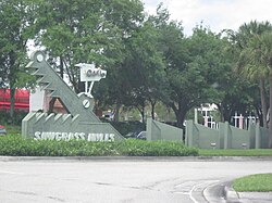 ป้ายรูปจระเข้ที่มีข้อความ "Sawgrass Mills" ตัวพิมพ์ใหญ่ทั้งหมดเป็นตัวเอียงอยู่ด้านบน และมีป้ายรูปนกที่มีข้อความ "The Oasis" ตัวพิมพ์ใหญ่ทั้งหมดเช่นกันอยู่ด้านบน