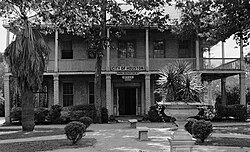 บ้านอิฐสองชั้นหลังคาทรงจั่ว มีระเบียงรอบบ้านที่สวยงาม และระเบียงชั้นสองที่มีเสา เสาสี่เหลี่ยมบนระเบียงชั้นล่างทำจากอิฐ