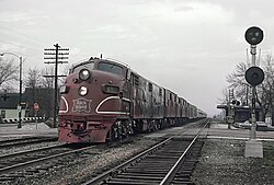 รถไฟหัวจักรไฟฟ้าสีแดงกำลังวิ่งเข้ามาหาผู้ดูจากเลนซ้ายในถนนที่ขับชิดขวา ตรงข้ามกับผู้ดู เสาโทรศัพท์ ป้ายหยุด และทางข้ามทางรถไฟสามารถมองเห็นได้ในระยะไกล