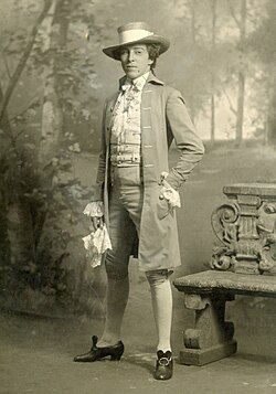 ภาพถ่ายขาวดำเต็มตัวของนักแสดงหญิงบนเวทีในชุดแต่งกายที่ประณีต