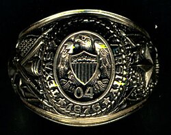 ภาพระยะใกล้ของแหวนทองคำ ด้านบนมีคำว่า Texas A&M University 1876 ล้อมรอบนกอินทรีที่อยู่บนโล่เหนือตัวเลข 0 และ 4 ด้านซ้ายมีดาวดวงใหญ่และต้นโอ๊ก ด้านขวามีปืนใหญ่ ดาบ และปืนไรเฟิล พร้อมธงสหรัฐอเมริกาและธงเท็กซัสไขว้กัน