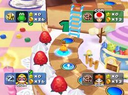 ภาพหน้าจอเกมรูปสี่เหลี่ยมผืนผ้าแนวนอน ซึ่งเป็นภาพดิจิทัลของกระดานเกมธีมขนมหวาน เส้นทางที่ทำจากคุกกี้ลายตารางหมากรุกวางอยู่บนเค้กสีขาวขนาดใหญ่ที่ตกแต่งด้วยน้ำตาลไอซิ่งและสตรอว์เบอร์รีขนาดยักษ์ เจ้าหญิงในชุดสีส้มยืนอยู่บนวงกลมสีฟ้าหลายวงเรียงกันตามเส้นทาง ข้อมูลตัวละครพร้อมลำดับขั้นและตัวนับดาวและเหรียญปรากฏอยู่ที่มุมแต่ละมุมของหน้าจอ