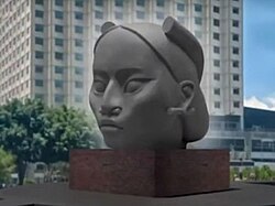 รูปปั้นศีรษะขนาดใหญ่ของหญิงสาวในรูปแบบที่เรียบง่าย ตั้งอยู่บนฐานสี่เหลี่ยมผืนผ้าในพื้นที่กลางแจ้งของเมือง