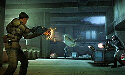 ในภาพด้านหน้า หญิงสาวจากเกม Half-Life 2: Deathmatch ถืออาวุธที่เรืองแสงสีส้มอยู่ ส่วนในฉากหลัง โถส้วมที่ชำรุดถูกยกขึ้นกลางอากาศด้วยปืน และเล็งไปที่ศัตรูที่ถือปืนอยู่เช่นกัน